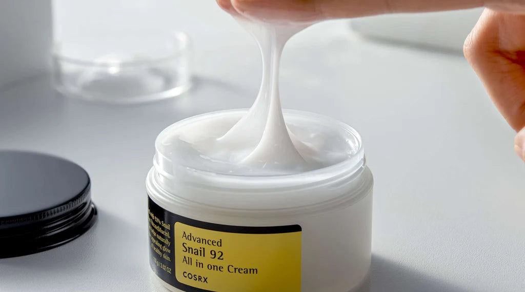Open jar of Cosrx Snail 92 cream moisturizer with creamy texture being lifted by fingers